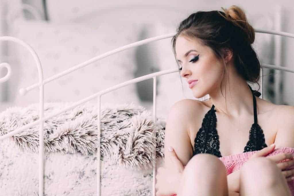A dark haired smiling woman in a black lacy bralette sitting next to her bed