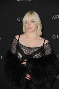 Billie Eilish wears lace-trimmed ivory over a sheer top with a fur jacket around her arms having red nails and a ruby necklace in an event place