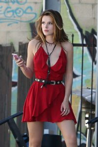 Bella Thorne wears a red spaghetti strap mini dress with a black belt and holds a smartphone in her right hand near a graffiti wall