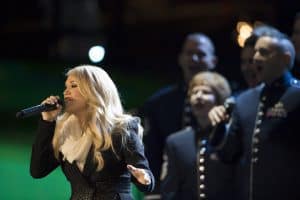 Carrie Underwood wears a black long-sleeve coat with a snowflake-like white speckled pattern and a cream scarf wrapped around her neck holding a black microphone while singing at a concert