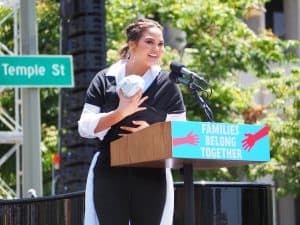 Chrissy Teigen wears a white long-sleeved blouse and black and white pants with a black baby wrap to carry her baby while speaking at L.A. Families Belong Together March