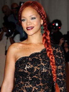 Rihanna with red braided hair and green dangling earrings wears a black lace evening gown at an event