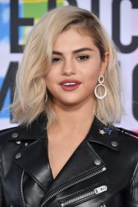 Selena Gomez with blond hair wears a black leather jacket and large diamond earrings while being photographed at the award show