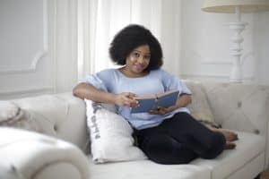 A woman wearing a light blue blouse and black skinny jeans holds a book while seated on a white couch in a living room