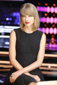 Taylor Swift wears a black mini dress with clear beads while seated on a chair in a well-lit studio