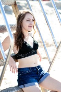 A woman in a black bralette and blue denim shorts seated on stainless stairs on the beach