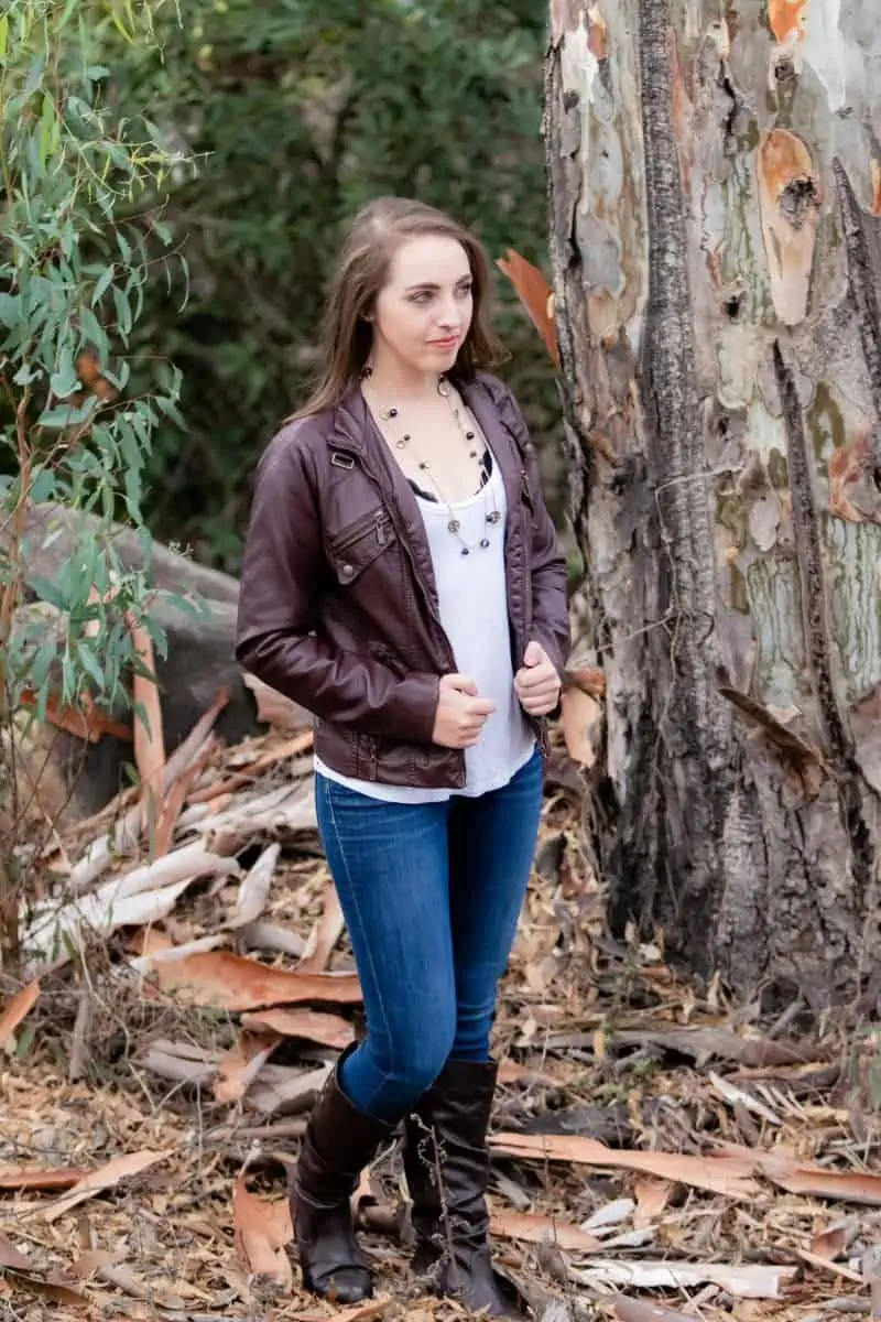 A woman in a brown leather jacket and white blouse wears blue jeans and black boots standing in the woods