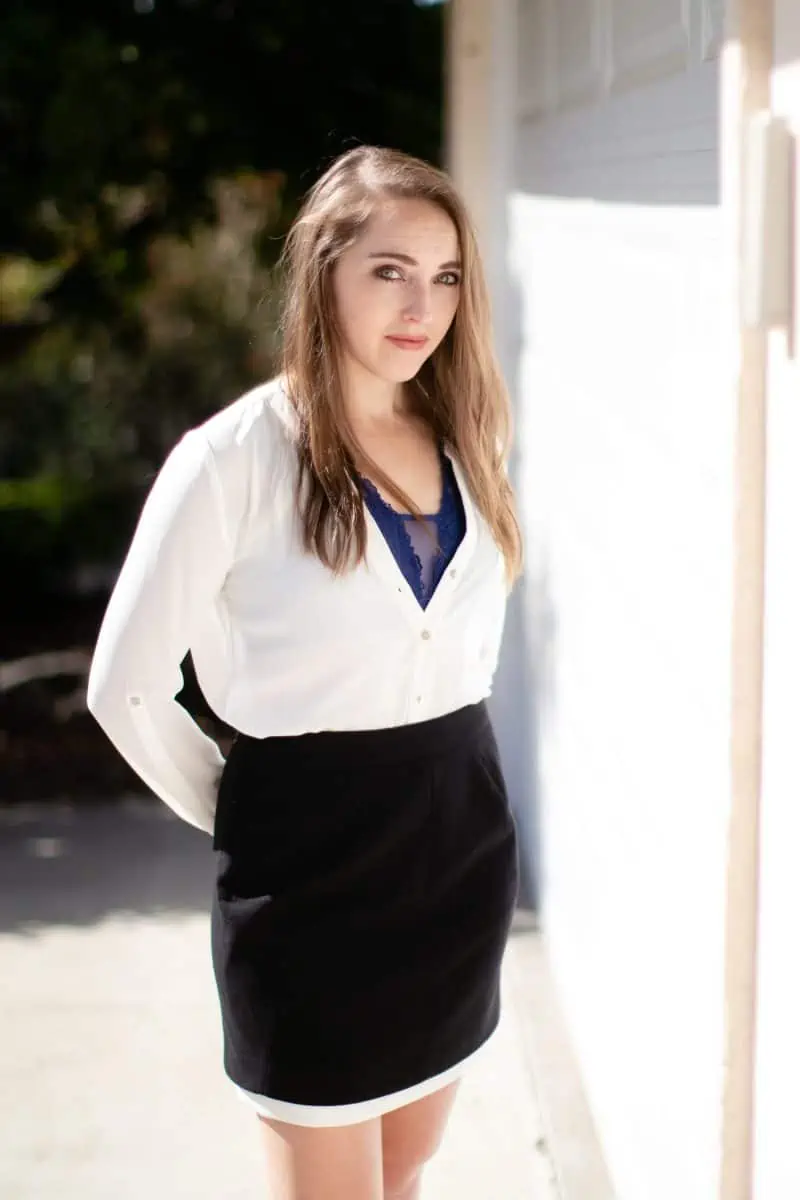 A woman in a blue bralette with a white long sleeves blouse and black mini skirt stands near a white wall on a sunny day