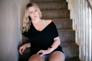 A woman wearing a black one off shoulder top and denim shorts seated on the stairs