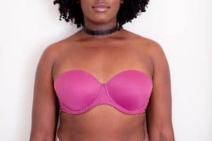 A pink strapless underwire bra and a black choker are worn by a woman standing near a white wall