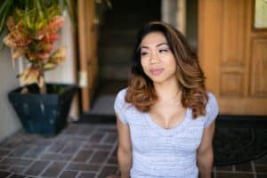 A woman with blond curly hair wears a gray v-neck shirt seated outside the house