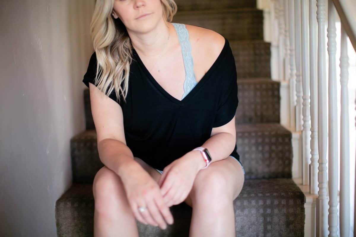 A woman in a black v-neck blouse on top of a blue bralette wearing denim shorts is seated on the stairs