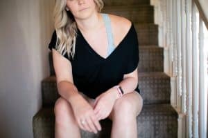 A woman in a black v-neck blouse on top of a blue bralette wearing denim shorts is seated on the stairs