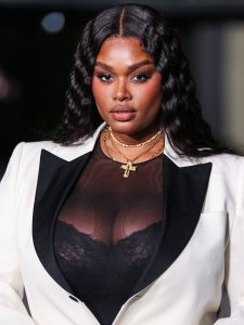 Precious Lee wearing a white tuxedo jacket and a see through black top that showcases her lacy black bra