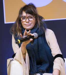 Lena Headey in a cream dress and a blue scarf wearing brown glasses at a conference
